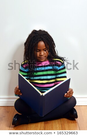 Foto d'archivio: Cute Black Hair Little Girl Reading Book By The Wall