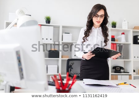 Stock foto: A Beautiful Young Girl Stands Near An Office Desk And Holds In Her Hands A Sheet For Notes And A Pen