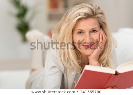 [[stock_photo]]: Smiling Blonde Woman Reading A Book While Lying On A Sofa In Her Appartment