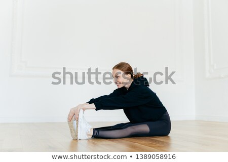 Foto d'archivio: Indoor Shot Of Smiling Redhead Woman Stretches Legs Focused Into Distance Touches Feet Wears Whit