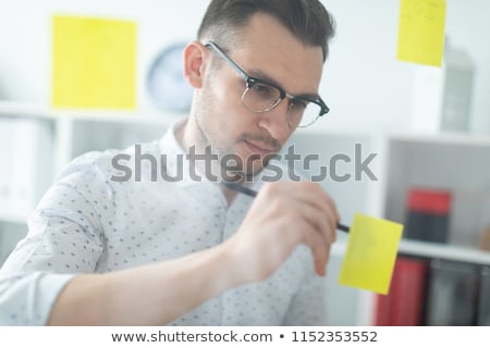 Stok fotoğraf: A Young Man In Glasses Stands Near A Board With Stickers And Holds A Glass Of Coffee In His Hands