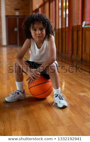 Foto d'archivio: Front View Of A Mixed Race Schoolboy Sitting On Basketball And Looking At Camera In Basketball Court