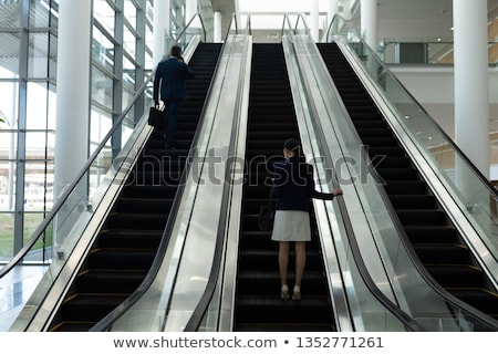 Foto d'archivio: Rear View Of Businessman And Businesswoman Moving Upwards On Escalator In Office Lobby