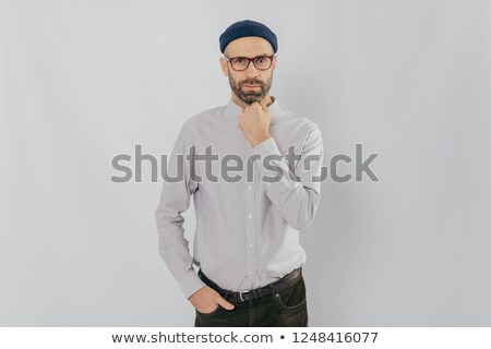 Foto d'archivio: Waist Up Shot Of Serious Man Keeps One Hand Under Chin Other In Pocket Has Stubble Wears Hat Whi