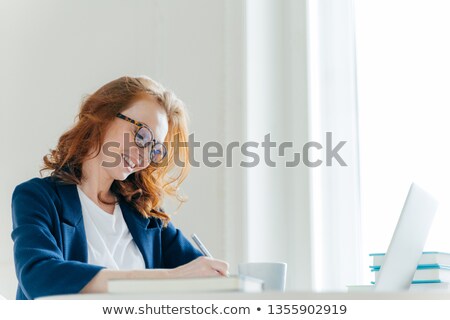 Foto stock: Indoor Shot Of Pleased Cheerful Famale With Ginger Hair Signs Contract Sits At Cabinet In Front Of