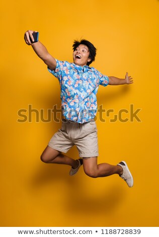 Foto d'archivio: Emotional Young Asian Man Jumping Isolated Over Yellow Background Take A Selfie By Mobile Phone
