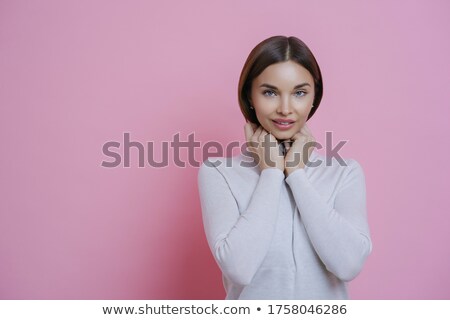 Stockfoto: Photo Of Pleased Dark Haired Woman Looks With Shy Expression At Camera Keeps Hand Under Chin Wears