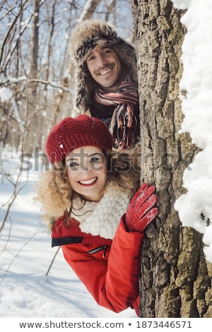 Stock fotó: Playful Couple In The Snow Hiding Behind A Tree Trunk