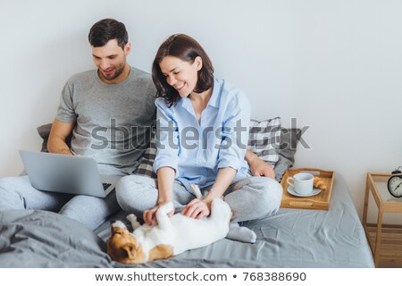 Foto d'archivio: Cheerful Brunette Young Female Plays With Her Favourite Dog In Bed While Her Husband Surfes Social N