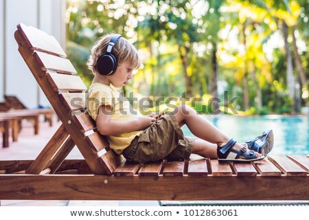 Stockfoto: A Cute Little Boy Is Using A Smartphone Lying On A Deckchair By The Pool Primary Education Friends