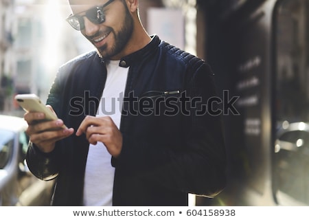 Foto d'archivio: Smiling Bearded Young Man In Glasses Using Smartphone Outdoors