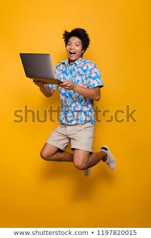 Stockfoto: Happy Young Asian Man Standing Isolated Over Yellow Background Using Laptop Computer Holding Debit C