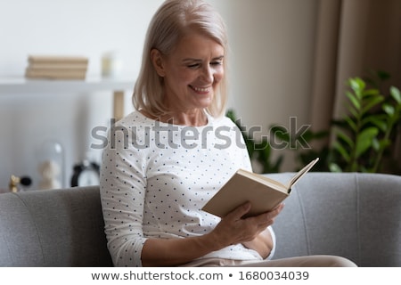 Foto stock: Concentrated Blonde Lady Sitting Indoors Reading Book