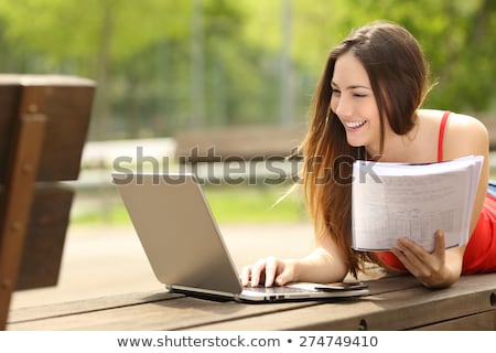 Stockfoto: Young Woman Lies Outdoors In Park Reading Book Using Laptop Computer