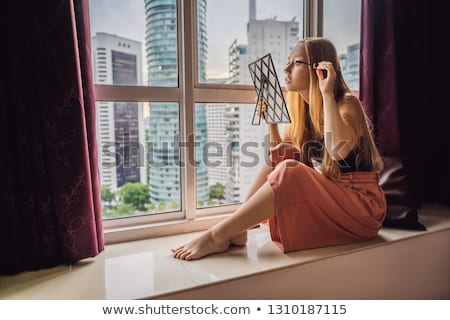 Stockfoto: Young Woman Does Makeup Sitting By The Window With A Panoramic View Of The Skyscrapers And The Big C
