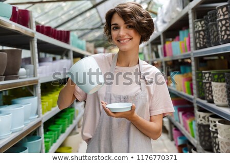 [[stock_photo]]: Woman Gardener Standing In Greenhouse Choosing Vase Pot For Plants