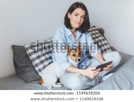 Stockfoto: Dreamy Brunette Female Model In Pyjamas Sits Crossed Legs On Comfortable Bed In Bedroom Holds Moder