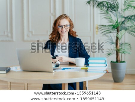 Stockfoto: Indoor Shot Of Red Haired Female Economist Sends Text Message To Colleague Holds Contemporary Smart