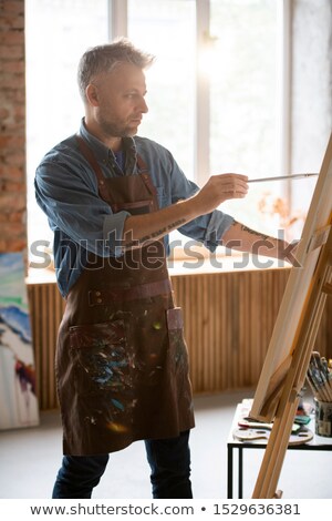 Foto stock: Serious Man With Paintbrush Looking At Picture On Easel While Painting