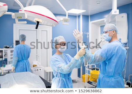 Stok fotoğraf: Young Assistant Putting Gloves On Hands Of Surgeon While Preparing For Operation
