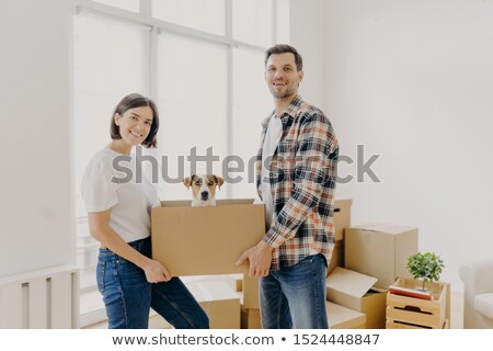 Stockfoto: Happy Family Couple Hold Carton Box With Small Puppy Stand Indoor Against Big Window Glad To Becom