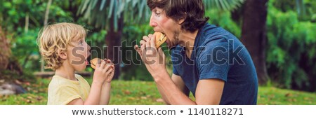 Foto d'archivio: Portrait Of A Young Father And His Son Enjoying A Hamburger In A Park And Smiling
