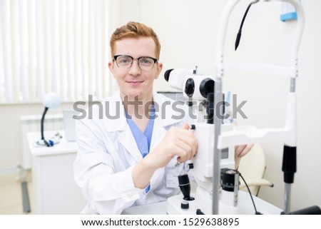 Stock photo: Happy Young Intern In Whitecoat And Eyeglasses Looking At You By Workplace