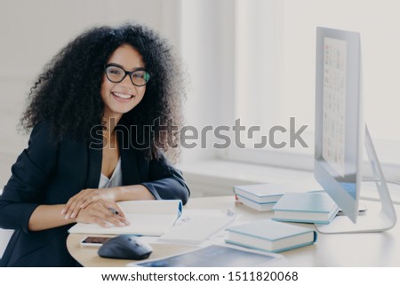 Stock photo: Glad Curly Office Worker Watches Webinar On Business Topic Writes Down Notes With Pen Smiles Posit