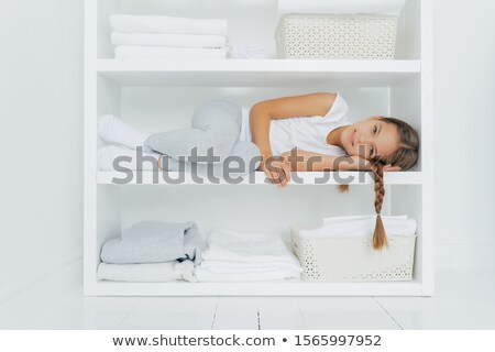 Stockfoto: Horizontal Shot Of Restful Girl With Pigtail Lies On White Console Shelf Dressed In Casual Clothes