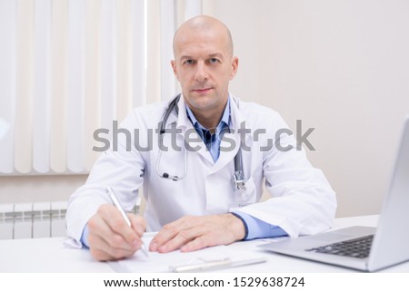 Zdjęcia stock: Bald Middle Aged Confident Doctor In Whitecoat Sitting By Workplace