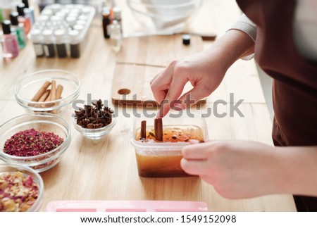 Foto d'archivio: Hands Of Young Woman Putting Cinnamon Sticks Into Liquid Soap Mass In Container