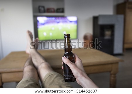 Foto d'archivio: Young Man Holding Soccer Ball And Beer And Watching Tv Translati