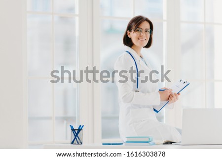 [[stock_photo]]: Skilled Female Physician Writes In Clipboard Poses At Workplace With Laptop Computer Professional