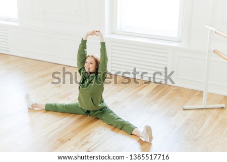 Foto stock: Strong Flexible Curly Red Haired Woman Does Side Leg Split Raises Hands Wants To Have Perfect Body