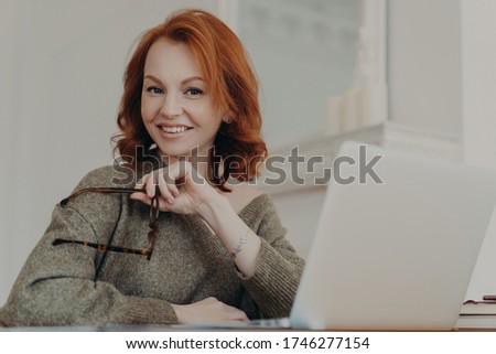 Stock photo: Skilled Successful Redhead Woman Holds Eyewear For Vision Protection Sits In Front Of Opened Laptop