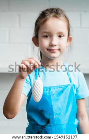 Stock photo: Cute Girl In Blue Apron Holding White Painted Handmade Decorative Firtree Cone