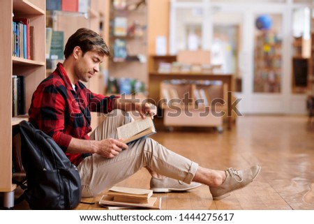 Foto d'archivio: Handsome Student In Casualwear Reading Book Or Manual In College Library