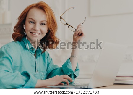 Foto stock: Glad Red Haired Woman In Blue Shirt Works On Laptop Uses Earphones For Watching Webinar Or Online