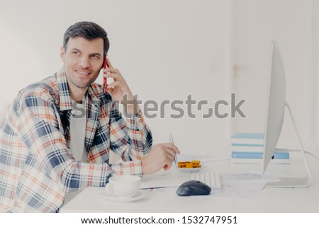 Сток-фото: Horizontal Shot Of Busy Male Freelancer Has Telephone Conversation Dressed In Checkered Shirt Writ