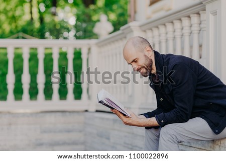 Сток-фото: People Hobby And Lifestyle Concept Postive Bald Man Reads Book With Enthusiasm Poses Outdoor Agai