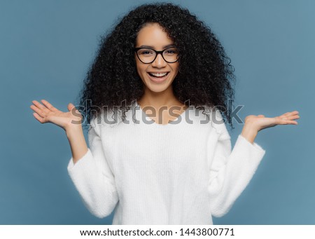 [[stock_photo]]: Positive Friendly Young Woman With Afro Appearance Spreads Palms Holds Invisible Object Has Charm