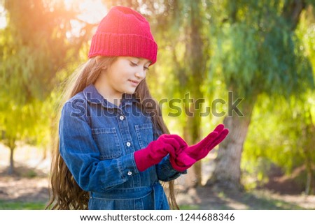 Stockfoto: Cute Mixed Race Young Girl Wearing Red Knit Cap Putting On Mitte