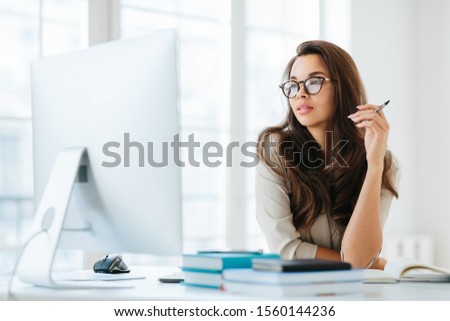Stock photo: Dark Haired Lady Focused Into Big Monitor Sits At Desktop Holds Pen And Writes Notes Wears Specta