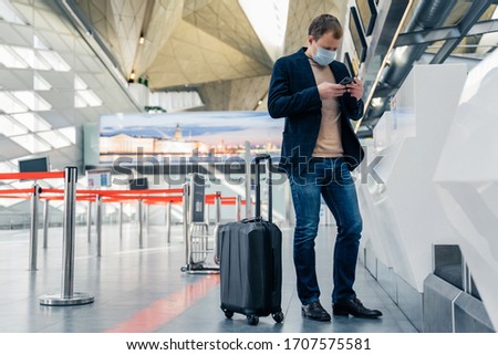 Stockfoto: Full Length Shot Of Man Passenger Stands Near Suitcase On Wheels In Airport Reads News Online On Cel