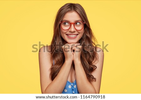 Stockfoto: Positive Young Caucasian Woman Keeps Hands Under Chin Has Dark Hair Toothy Smile Wears White Shir