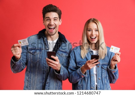 Stok fotoğraf: Excited Young Loving Couple In Jeans Denim Jackets Using Mobile Phones Isolated Over Red Background