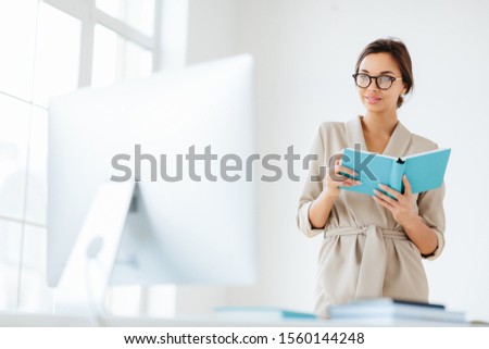 Zdjęcia stock: Young Dark Haired Woman In Elegant Wear Holds Blue Notepad Focused Into Monitor Ponders About Busin