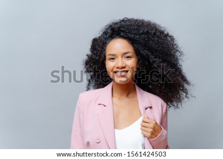 Сток-фото: Positive Curly Dark Skinned Woman With Luminous Hair Wears Formal Purple Jacket Poses Against Grey