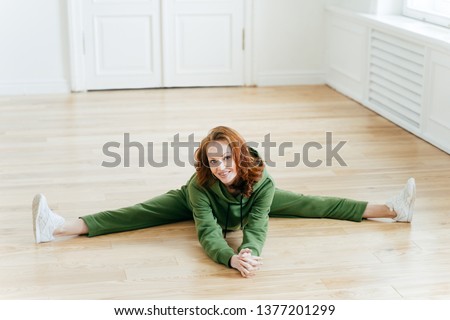 [[stock_photo]]: Horizontal Shot Of Positive Redhaired Woman Does Pilates Exercises Drssed In Green Tracksuit Has N