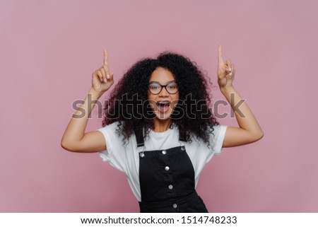 Stockfoto: Photo Of Happy Impressed Afro American Woman Points Both Index Finger Above Demonstrates Awesome Co